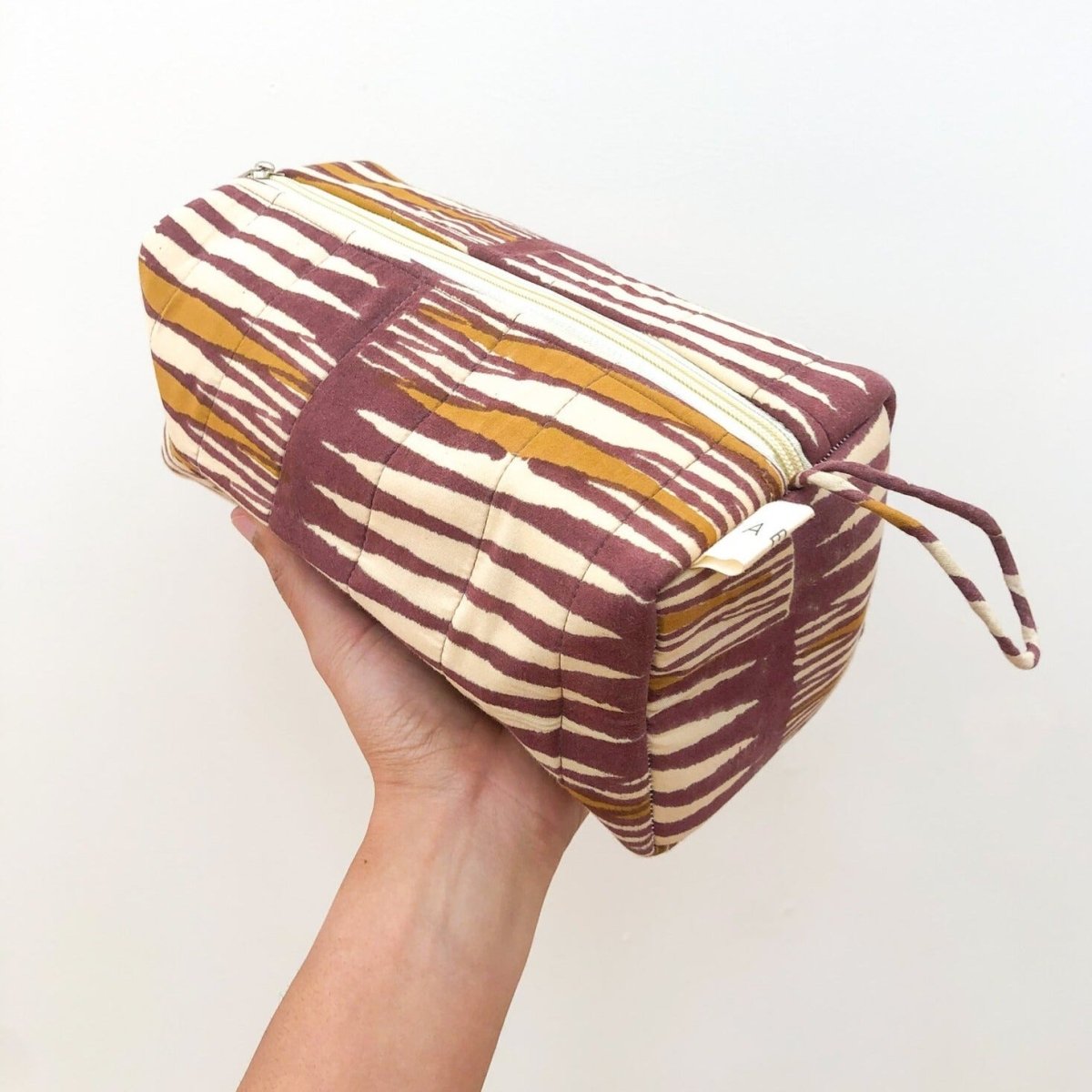 A person holding a maroon, mustard and cream patterned cosmetic bag. The Paloma Cosmetic Bag from Maelu is designed in Portland, Oregon and printed and made in India.