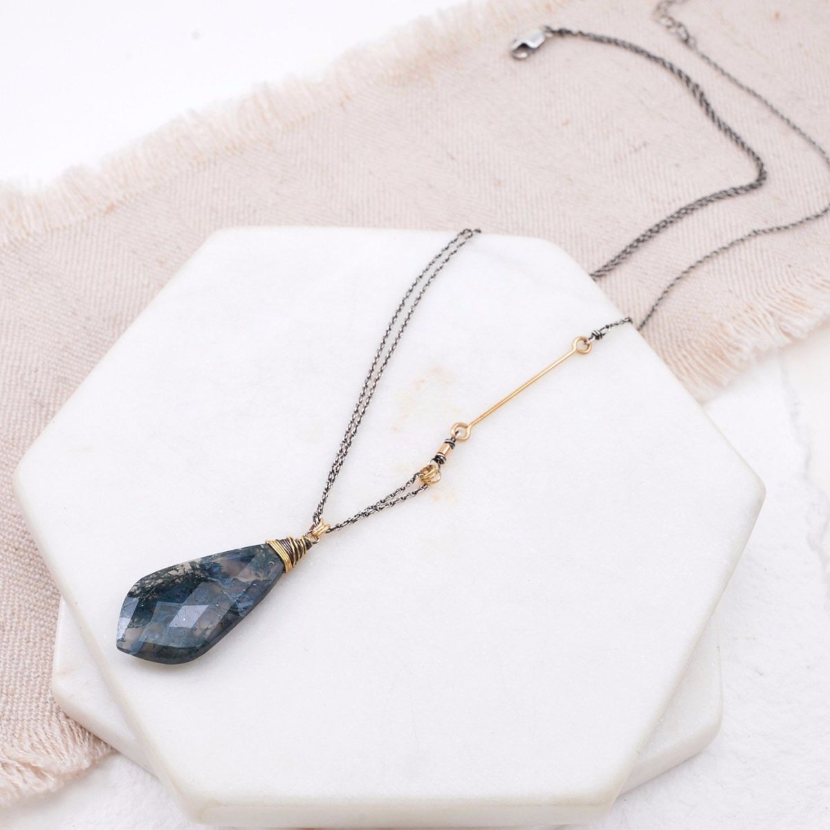 A blue, grey and black agate pendent hangs from a wrapping of gold-fill and sterling silver wire. Three tiny gold-fill hoops connect the pendant to a sterling silver chain with a gold filled accent bar. The Alloria Moss Agate Necklace is designed and handmade by Amy Olson in Portland, OR.