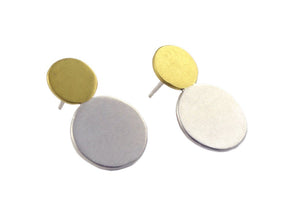 A pair of brass and silver earrings sit at an angle against a white background. The earrings are comprised of two circles– a smaller brass circle sits atop a medium silver circle. The Moonlet Post earrings are made by designer Natalie Joy in Portland, Oregon.