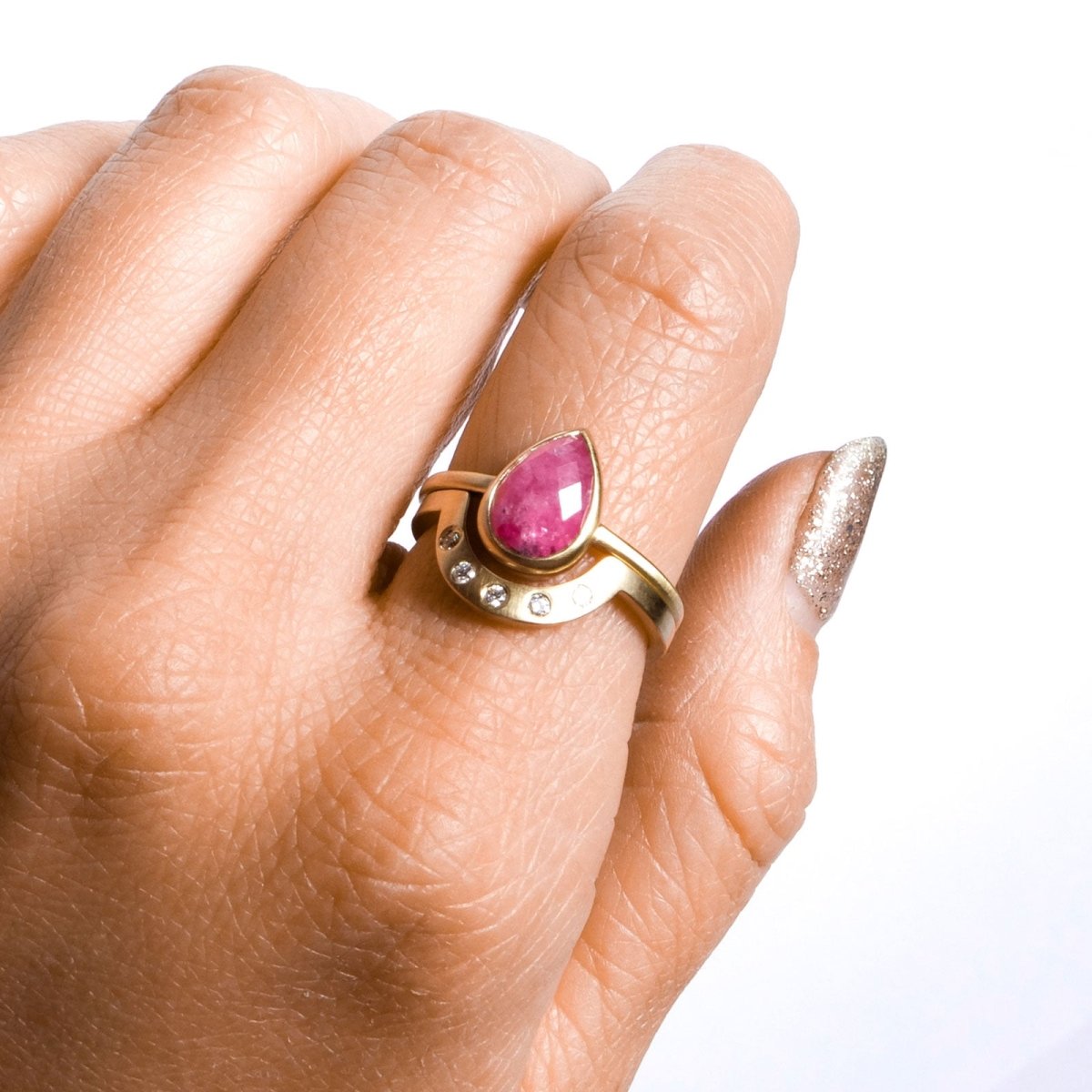 A model wears a conflict-free pear-shaped rose cut ruby set in a narrow 14k yellow gold band with a matte finish. Surrounding the ruby ring is an arch shaped yellow gold band with five lab grown white diamonds and a matte finish. The Pirum Ring and the White Diamond Cor Ring are designed by Betsy & Iya and handcrafted in Portland, Oregon.
