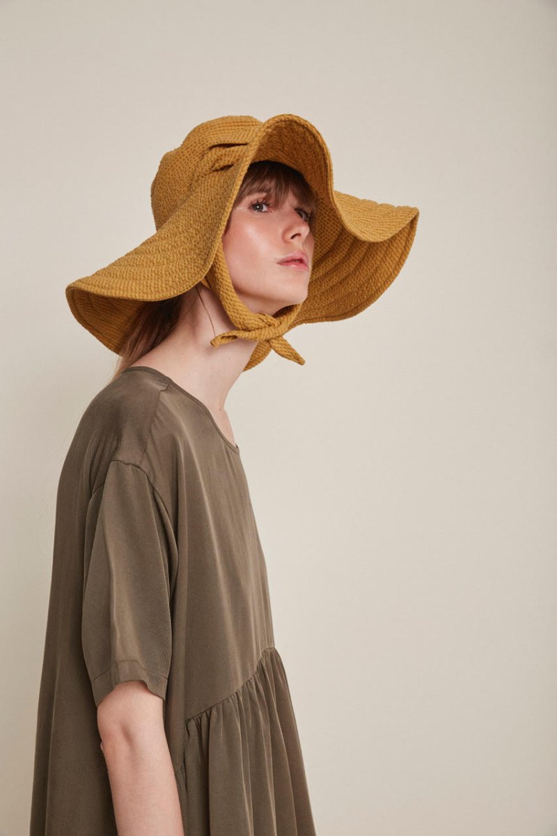 Red-haired woman wears a brown babydoll dress and yellow sun hat. The Drapey Dress in Kaki is from designer Rita Row.