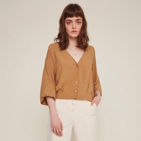 Red-haired woman wears a brown button-down blouse with loose sleeves and white pants. The Rosetta Blouse in Latte is from designer Rita Row.