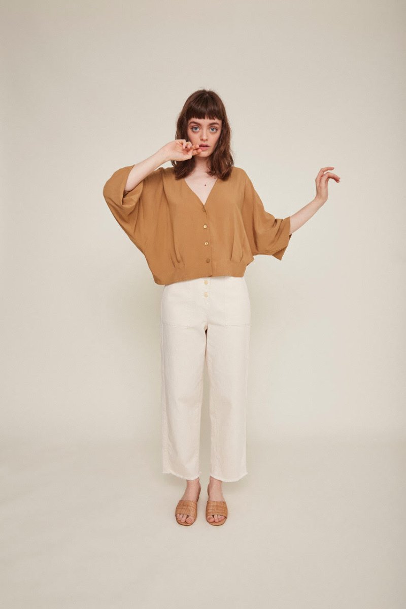 Red-haired woman wears a brown button-down blouse with loose sleeves and white pants. The Rosetta Blouse in Latte is from designer Rita Row.
