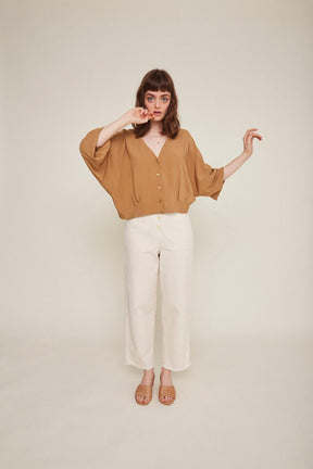 Red-haired woman wears a brown button-down blouse with loose sleeves and white pants. The Rosetta Blouse in Latte is from designer Rita Row.