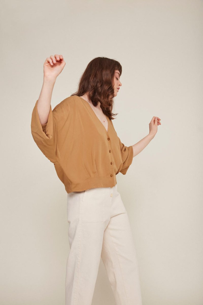Red-haired woman wears a brown button-down blouse with loose sleeves and white pants. She is looking away from the camera and dancing.  The Rosetta Blouse in Latte is from designer Rita Row.