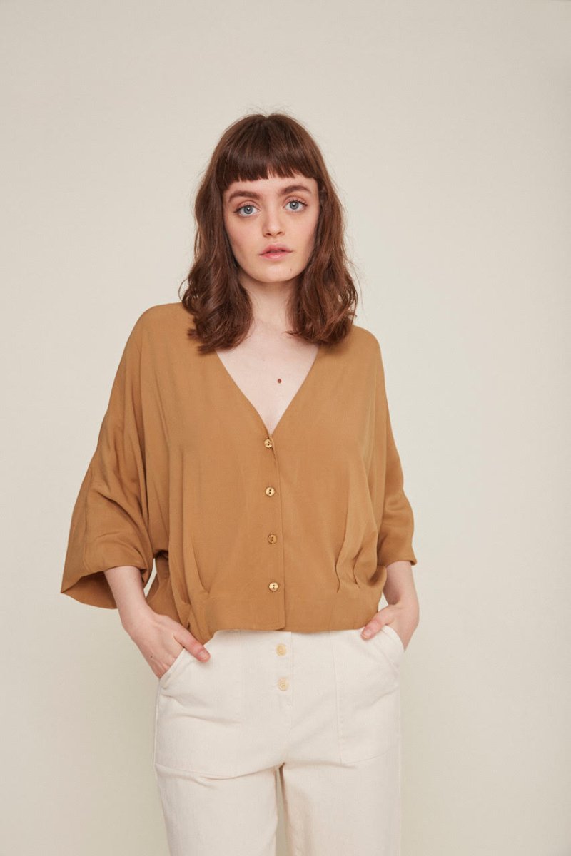 Red-haired woman wears a brown button-down blouse with loose sleeves and white pants. The Rosetta Blouse in Latte is from designer Rita Row.