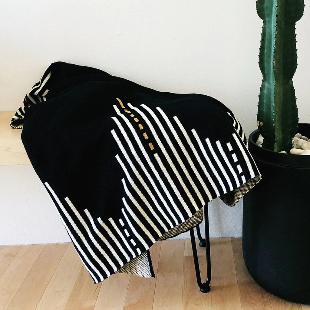 Black blanket with a white and gold striped pattern lays draped in a chair. The Reika Throw blanket from Seek and Swoon is designed in Portland, Oregon and made in USA.