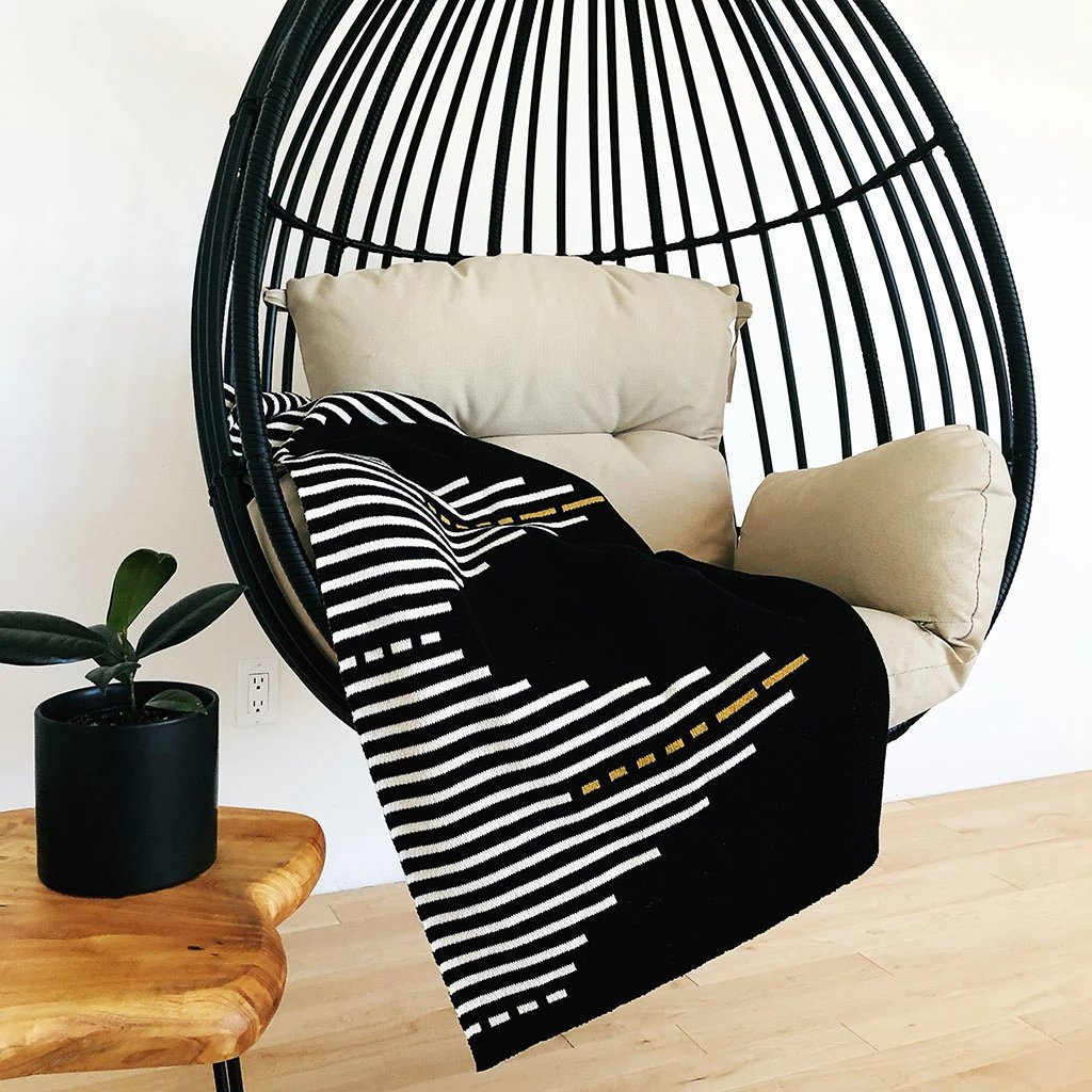 Black blanket with a white and gold striped pattern lays draped in a hanging chair. The Reika Throw blanket from Seek and Swoon is designed in Portland, Oregon and made in USA.