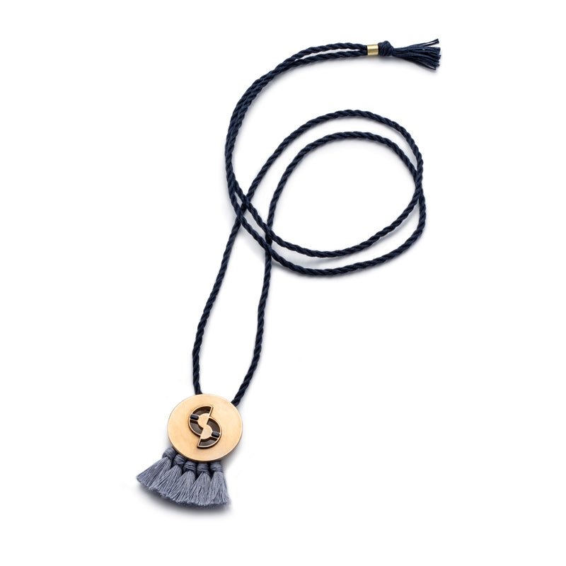 Circular, quarter-sized, cast-bronze pendant with curved cutouts, two rectangular, lapis lazuli inlays, and periwinkle cotton fringe, threaded with an adjustable, navy blue cotton rope. Hand-crafted in Portland, Oregon.