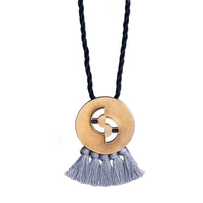 Circular, quarter-sized, cast-bronze pendant with curved cutouts, two rectangular, lapis lazuli inlays, and periwinkle cotton fringe, threaded with a navy blue cotton rope. Hand-crafted in Portland, Oregon.