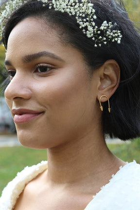 Woman wears white flowers in her hair and models "U" shaped brass earrings. The Which Way to Go Earrings are made by designer Lingua Nigra.