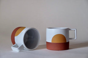 Two Sunrise Mugs against a white background. The mug on the left is laying on its side. The mug on the right is upright. The Sunrise Mugs from Wolf Ceramics have a white satin glaze and a golden yellow sunrise motif. Made in Portland, Oregon.